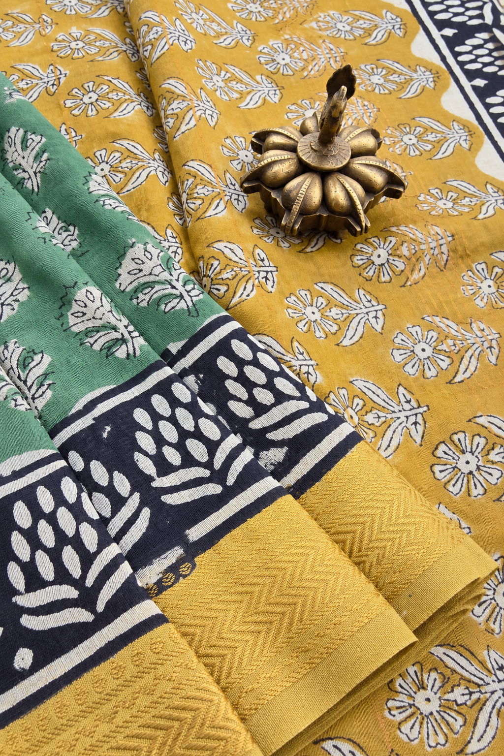 A folded block-printed fabric, likely a saree, featuring a mustard yellow body with white floral prints, a green border section with white floral prints, and a navy blue border with white leafy motifs and chevron patterns. A small brass oil lamp is placed on top of the fabric.