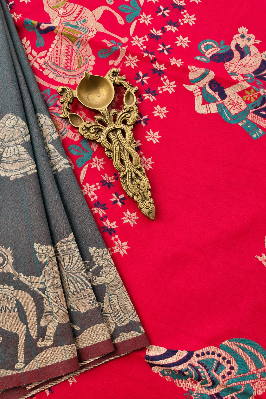A close-up of a richly patterned red and pink fabric, likely a saree, featuring traditional Indian motifs in gold, teal, and beige. A folded dark gray portion of the fabric with beige patterns is draped on the left, accented by an ornate antique gold metal oil lamp.